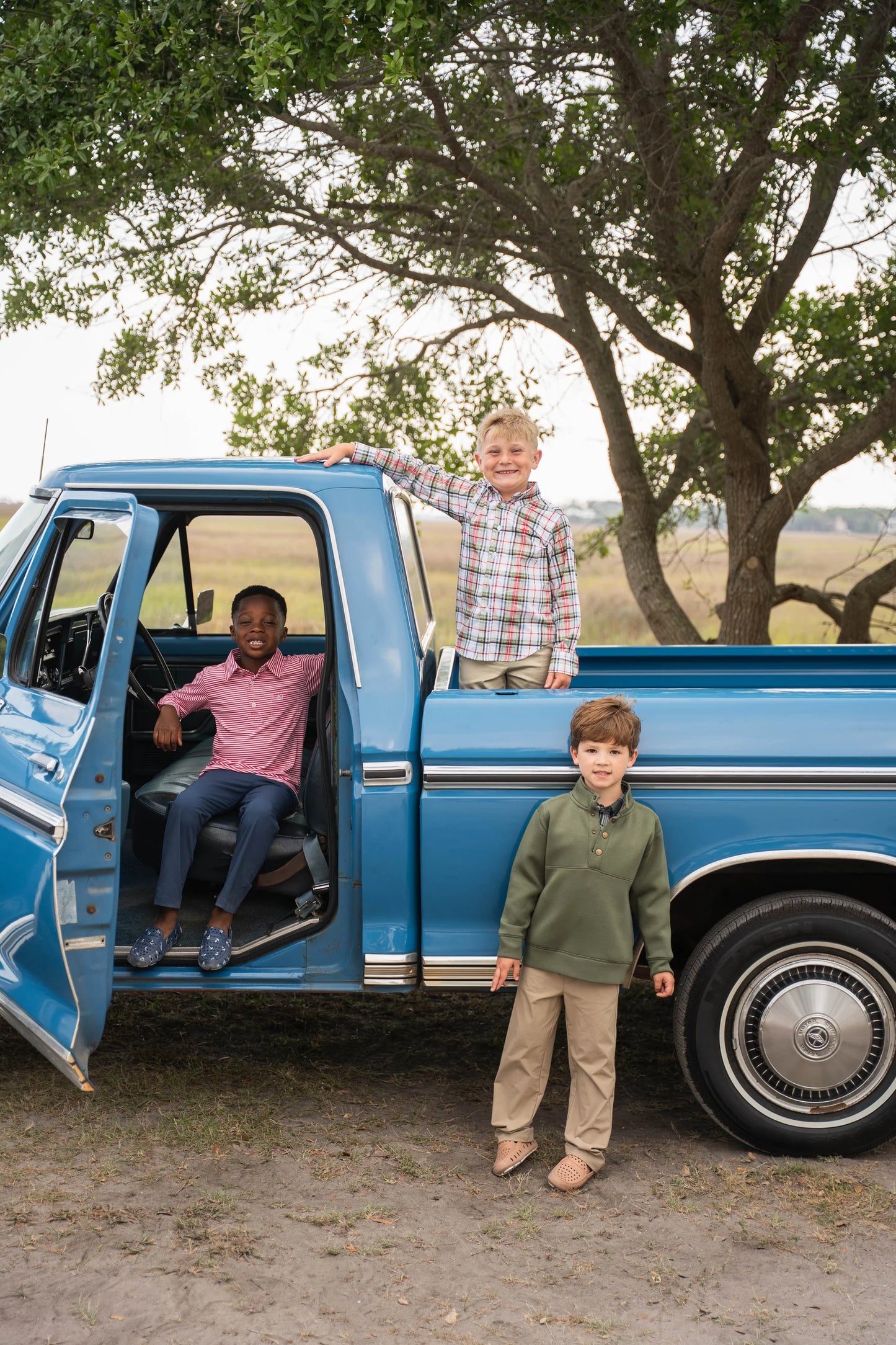 Boy in green Pierce Performance Pullover with performance pants standing in front of truck beside two boys in Oxford and other Saltwater Boys fall favorites, highlighting timeless fall outfit styles.