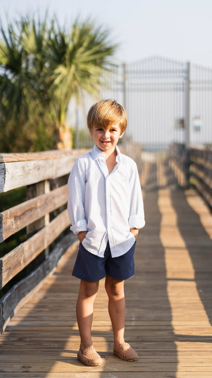 Boy walking on a beach boardwalk in the Carter Oxford Shirt in Seersucker by Saltwater Boys, showcasing its lightweight cotton and polished button-down style in a seaside setting