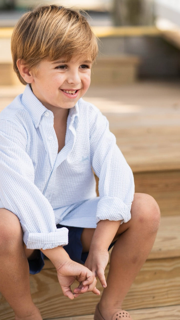 Boy wearing the Carter Oxford Shirt in Seersucker by Saltwater Boys, sitting on wooden beach steps, capturing the shirt’s relaxed yet dressed-up coastal style
