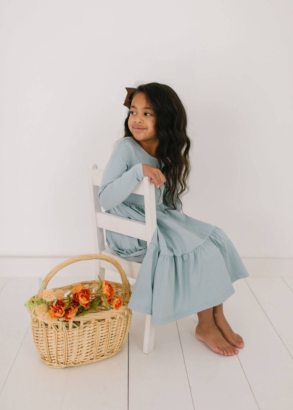 Toddler model sitting in the Gwendolyn Ruffle Dress in dusty blue, showing soft fabric and elegant ruffle accents.