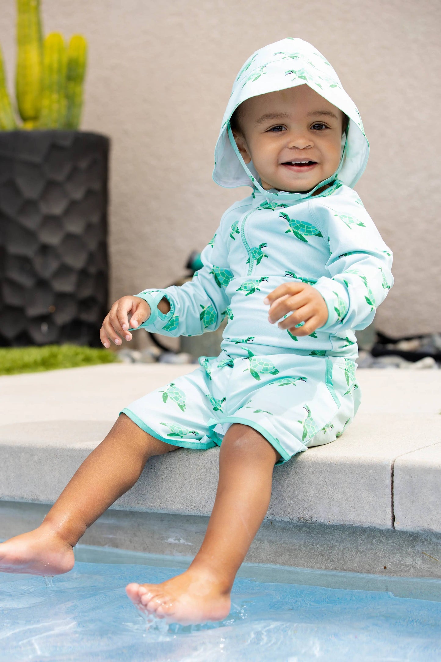 Child sitting at the edge of a pool wearing Sea Turtles sun hat, feet in water, enjoying the sun