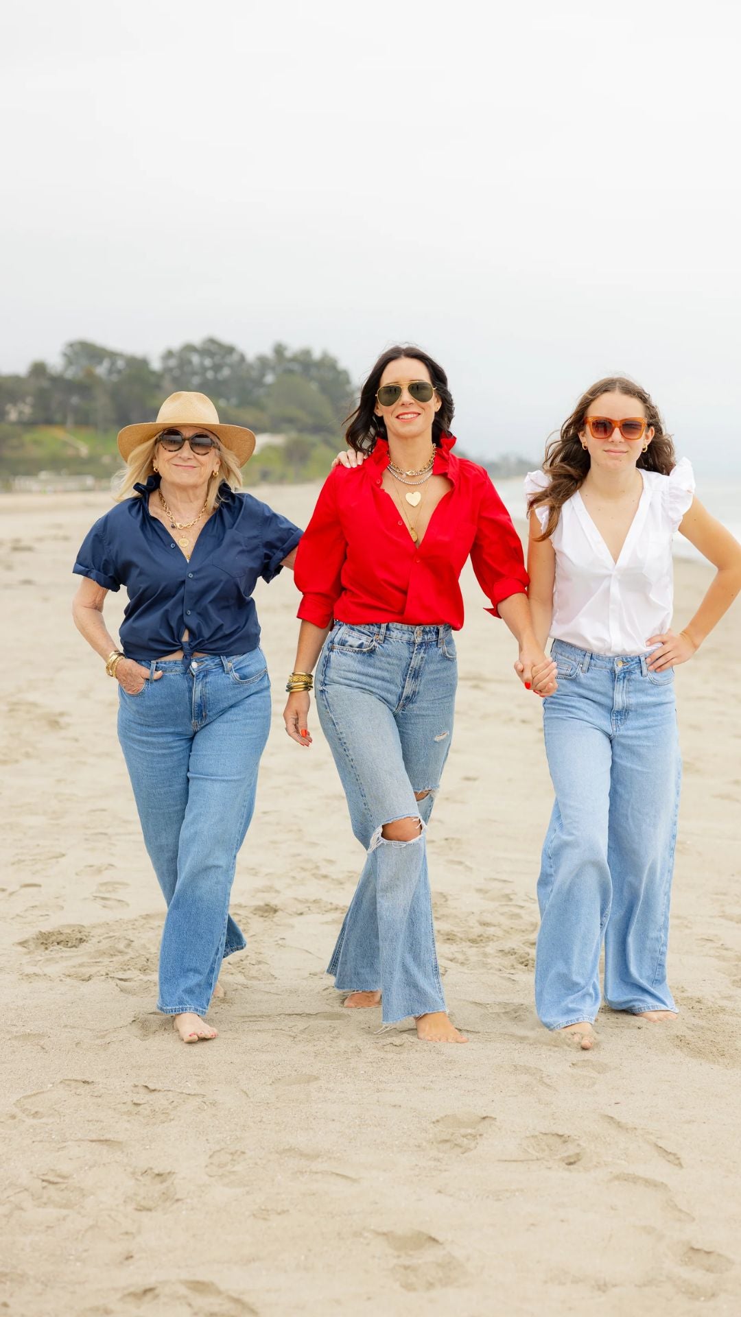 Three women of all ages walking side by side on the beach toward the camera—one wearing the KMJ Quarantini in navy, one in the Southern Lover in red (Kate, the designer), and one in the Olivia in white.