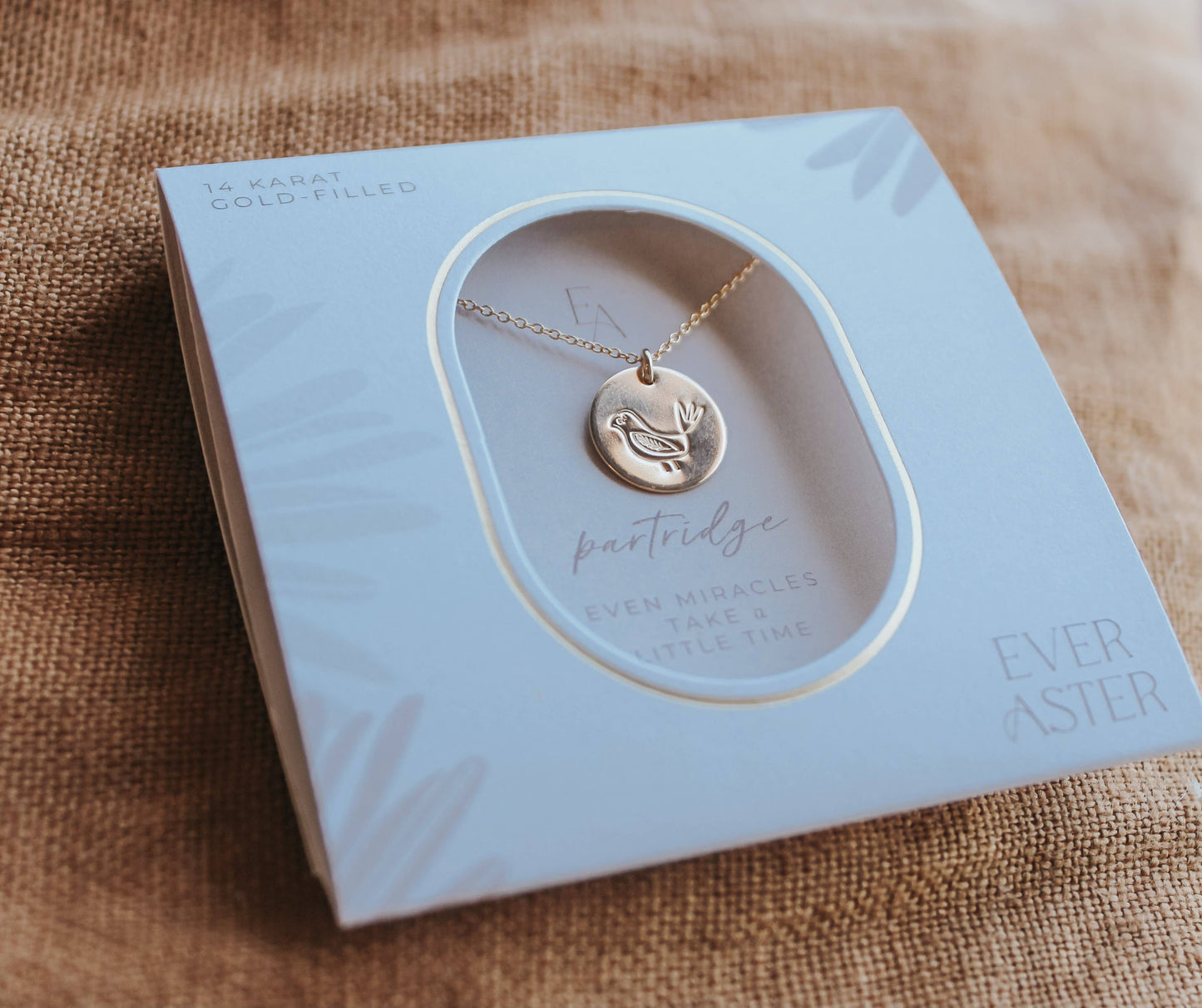 A flat lay of the Partridge Fertility Necklace by Ever Aster, crafted in gold-filled material. The delicate necklace is elegantly displayed in its packaging, with the heartwarming message