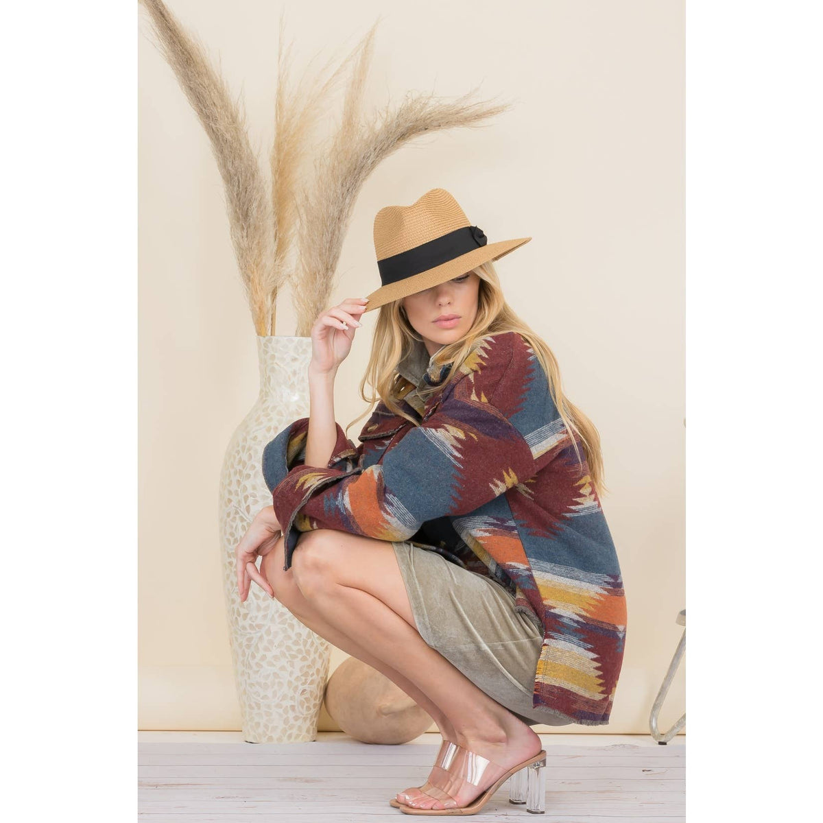 A woman wearing a colorful plaid fall jacket and beige shorts, crouching on a white wooden floor, with a tan wide-brimmed