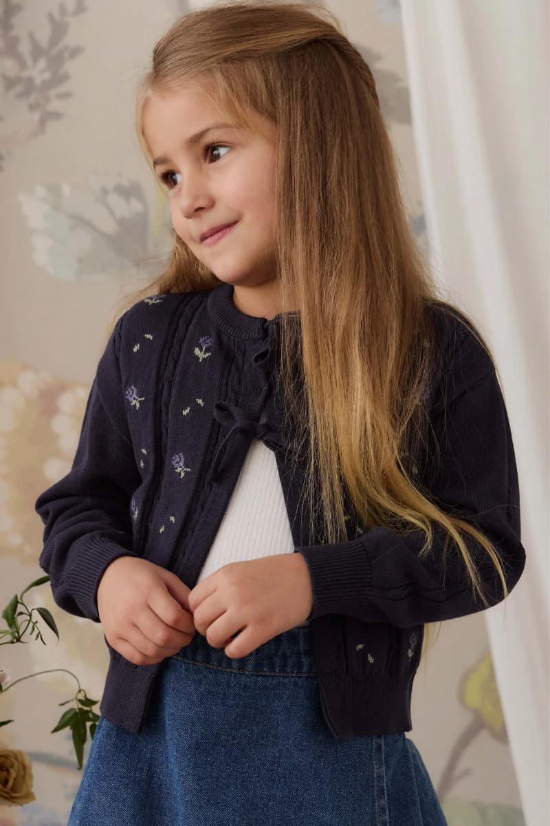 A young girl with long light brown hair wearing a deep purple knit cardigan with small purple floral details, layered over a white top and blue denim skirt, posing in an indoor setting with a floral wallpaper background.