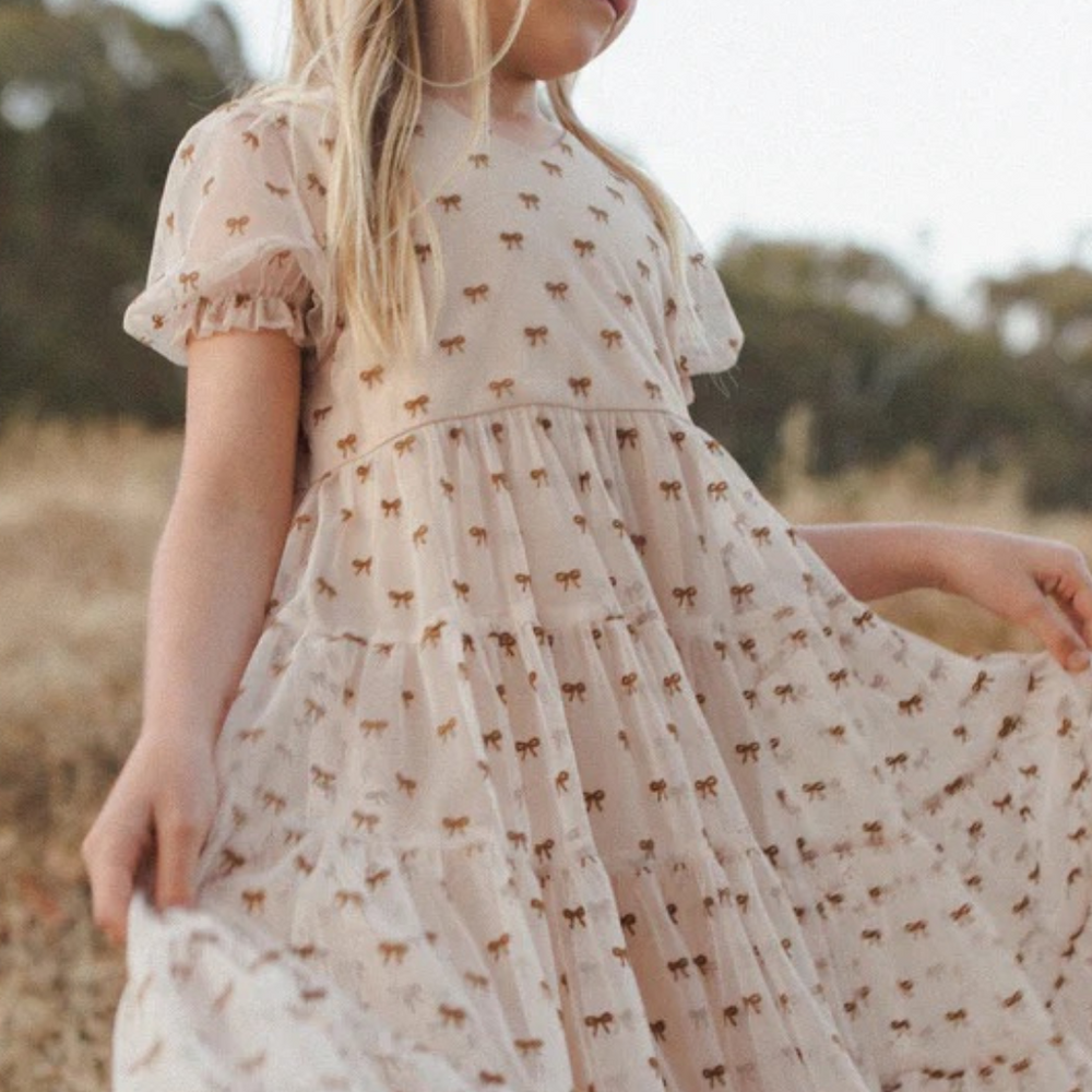 Girl twirling in The Dottie Dress by Noralee – A young girl twirls joyfully in The Dottie Dress by Noralee, showcasing its playful bow pattern and puff sleeves, perfect for special occasions and everyday style.