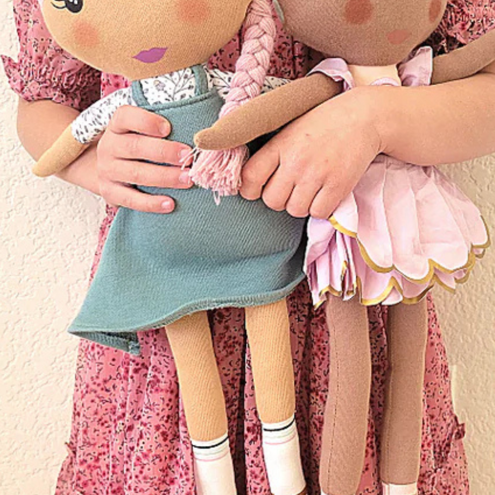 
                      
                        Up close of the Fair Kindness Doll next to another kindness doll, both being lovingly held by a child, highlighting their collectible nature.
                      
                    