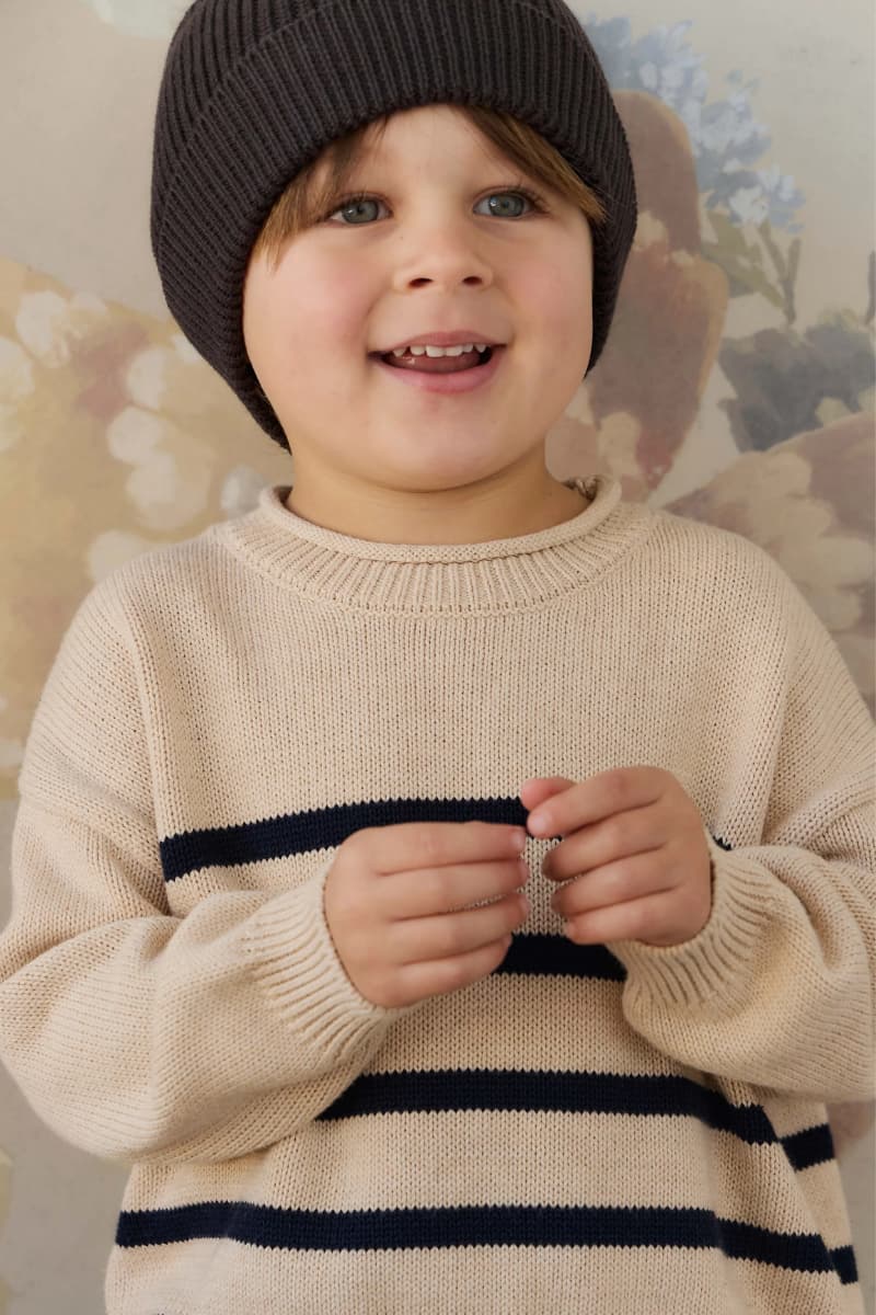 A young child wearing a cream knit sweater with navy horizontal stripes by Jamie Kay and a dark knit beanie, standing against a soft floral background. Available at Grace & Haven in Essex, CT or online at graceandhaven.co with free shipping always.