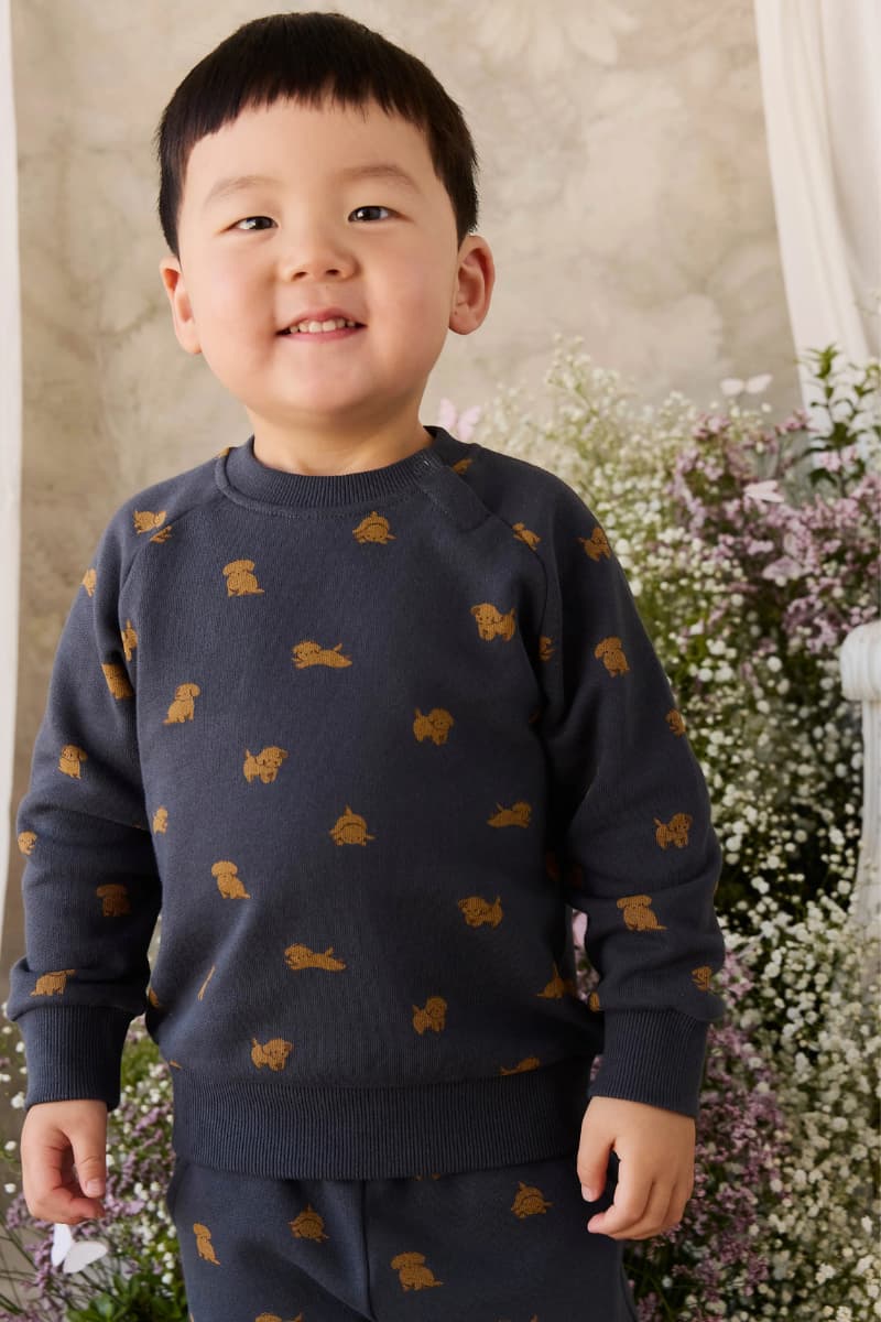 Child wearing a navy sweater with gold puppy dog prints by Jamie Kay in a floral setting. Available at Grace & Haven in Essex, CT or online at graceandhaven.co with free shipping always.