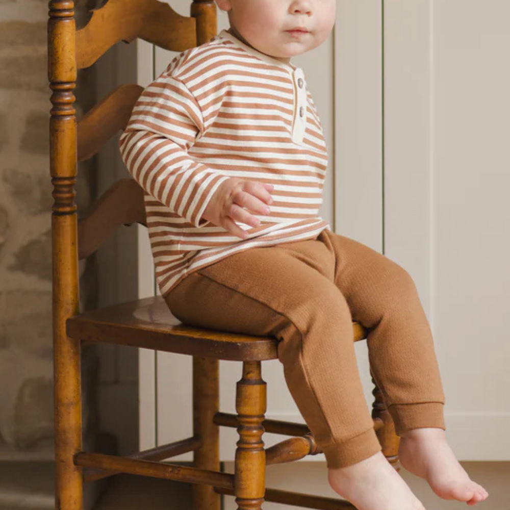 
                      
                        Toddler wearing cinnamon stripe Henley Tee, sitting and smiling on an antique kitchen chair.
                      
                    