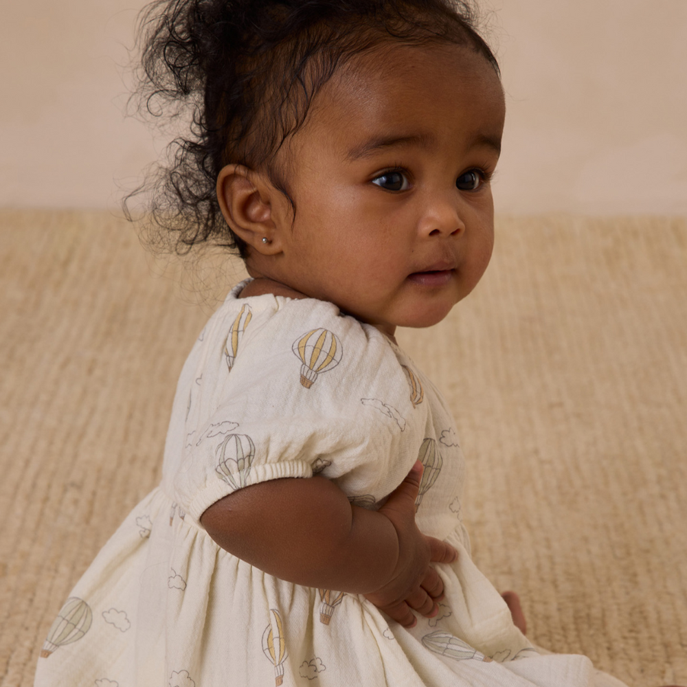 A charming side profile of a baby wearing the Quincy Mae Bella Dress, showcasing its whimsical hot air balloon print, delicate ruffle sleeves, and flowy silhouette, perfect for warm-weather adventures.