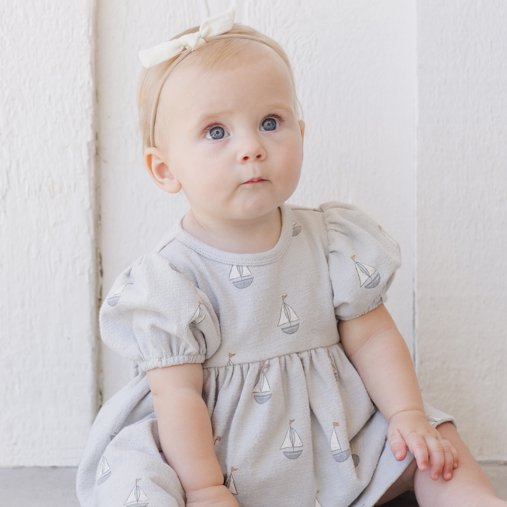 A sweet baby sitting in the Quincy Mae dress, showcasing its soft fabric, delicate design, and comfortable fit, perfect for warm-weather outings.