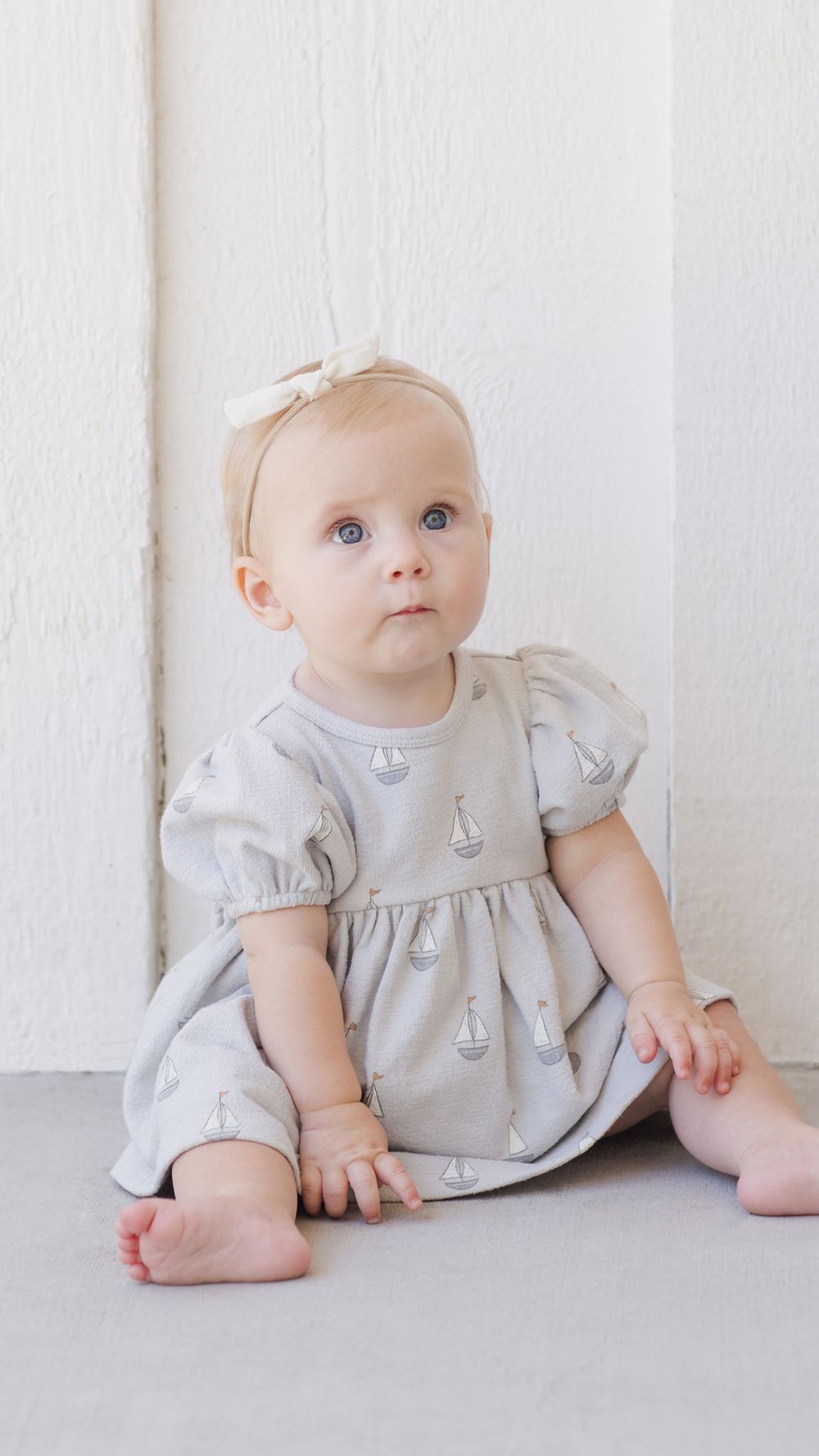 A sweet baby sitting in the Quincy Mae dress, showcasing its soft fabric, delicate design, and comfortable fit, perfect for warm-weather outings.