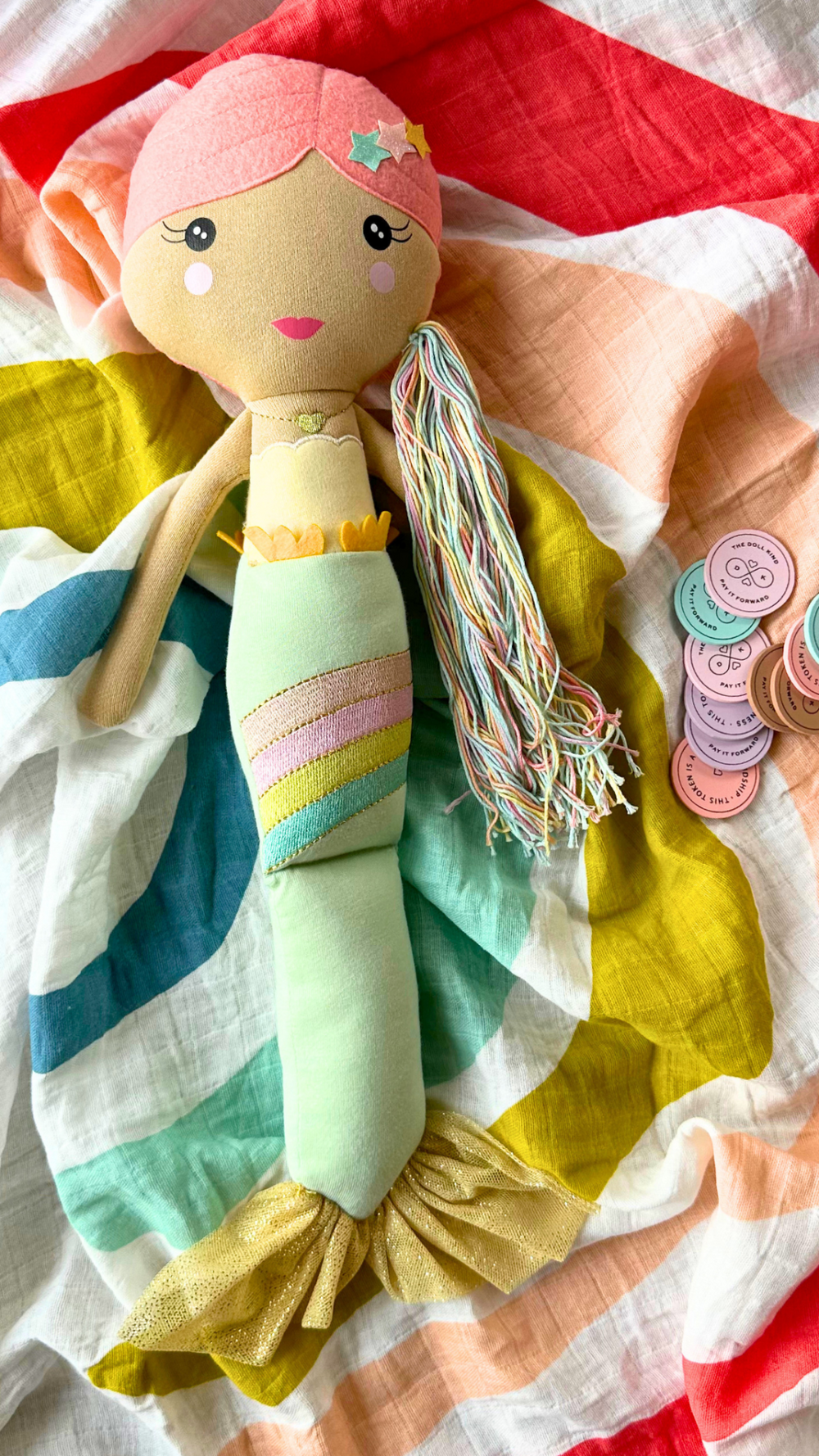 Beautiful arrangement featuring the enchanting Shine Mermaid Kindness Doll with kindness tokens, perfect for inspiring generosity and love, placed on a soft, colorful muslin swaddle.