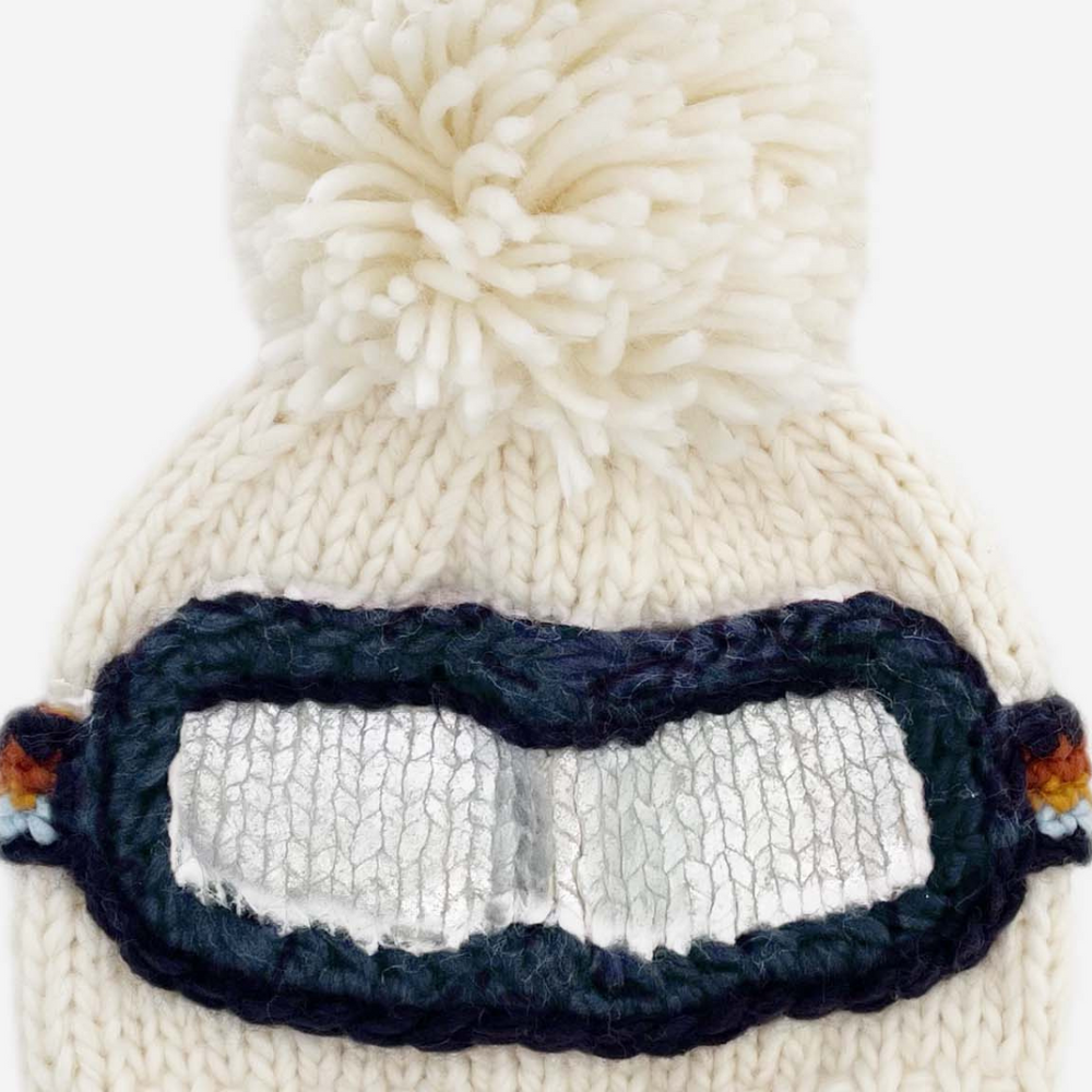 Front view of Ski Goggles Hat in cream with navy, cinnamon, mustard, and grey goggle band, topped with a playful pom-pom, perfect for winter adventures.