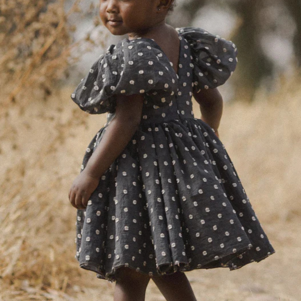 Model turned over her shoulder in a field, displaying the back of the Sophia Dress, which features a charming polka dot pattern and comfortable fit.