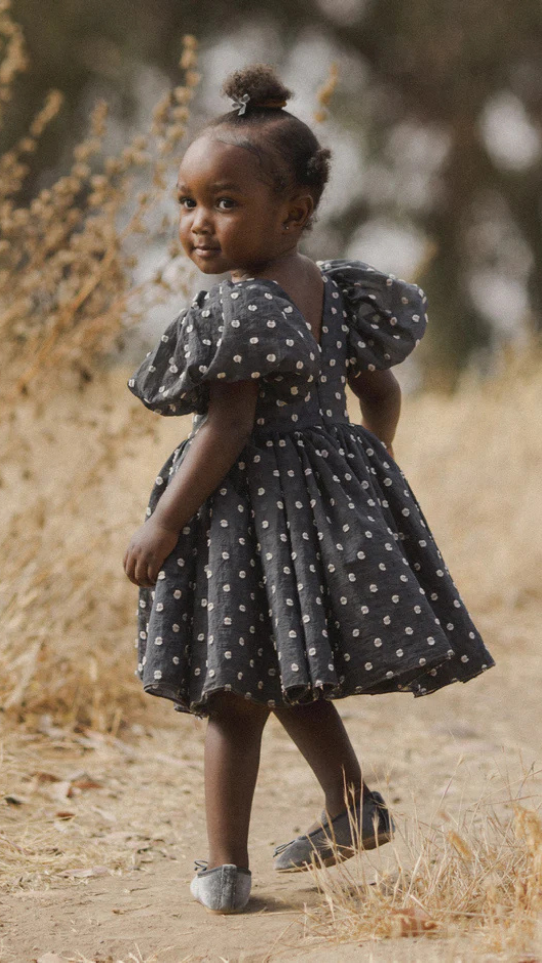 Model turned over her shoulder in a field, displaying the back of the Sophia Dress, which features a charming polka dot pattern and comfortable fit.