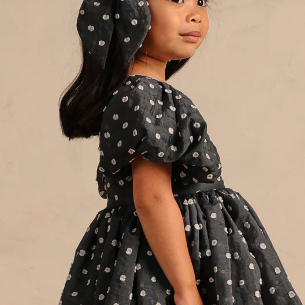 
                      
                        Side view of a model in the Sophia Dress, paired with an oversized bow in a studio setting, highlighting the playful elegance of the polka dot design.
                      
                    