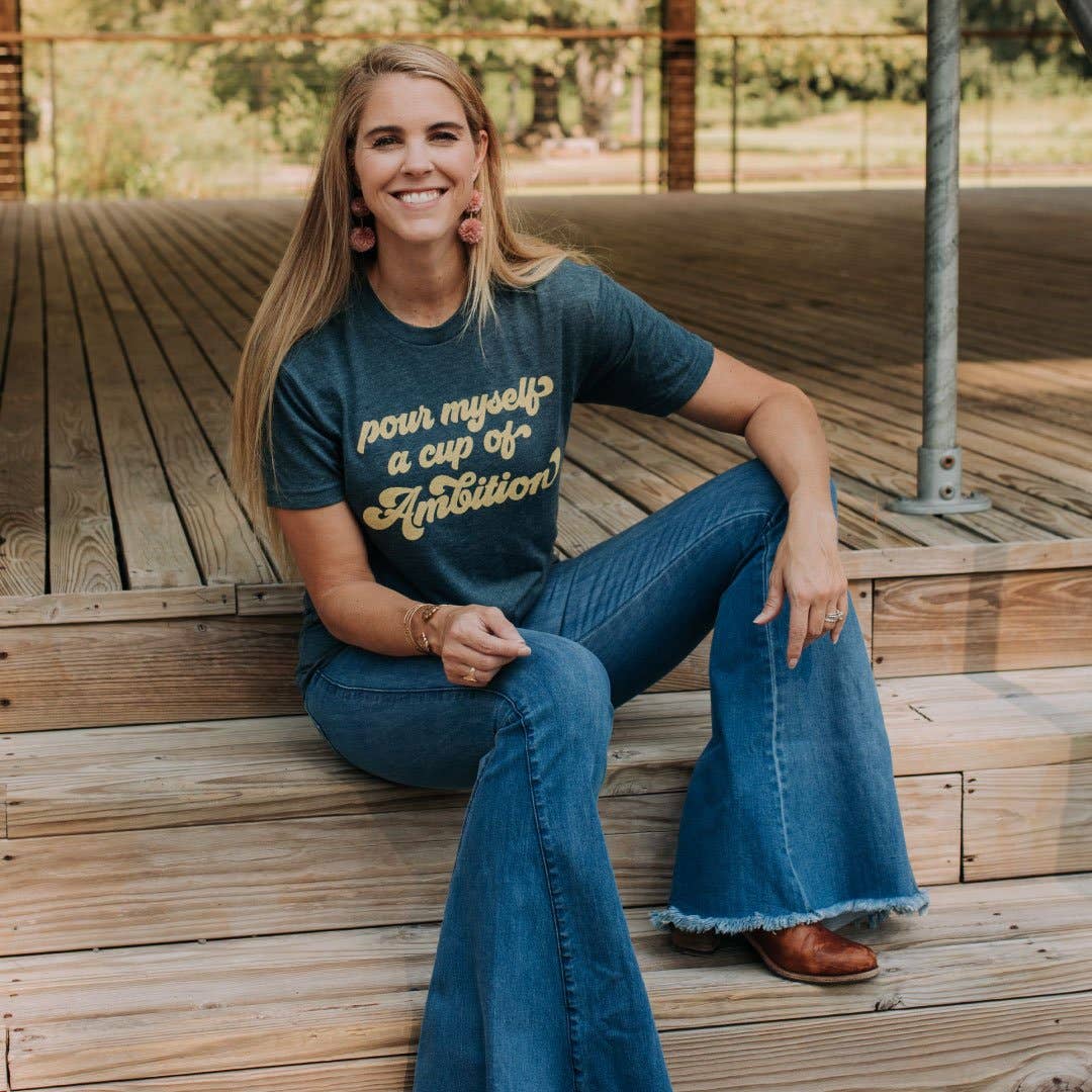 Woman sitting on wooden steps wearing a navy blue Pour Myself A Cup Of Ambition T-Shirt with beige text, blue jeans, and