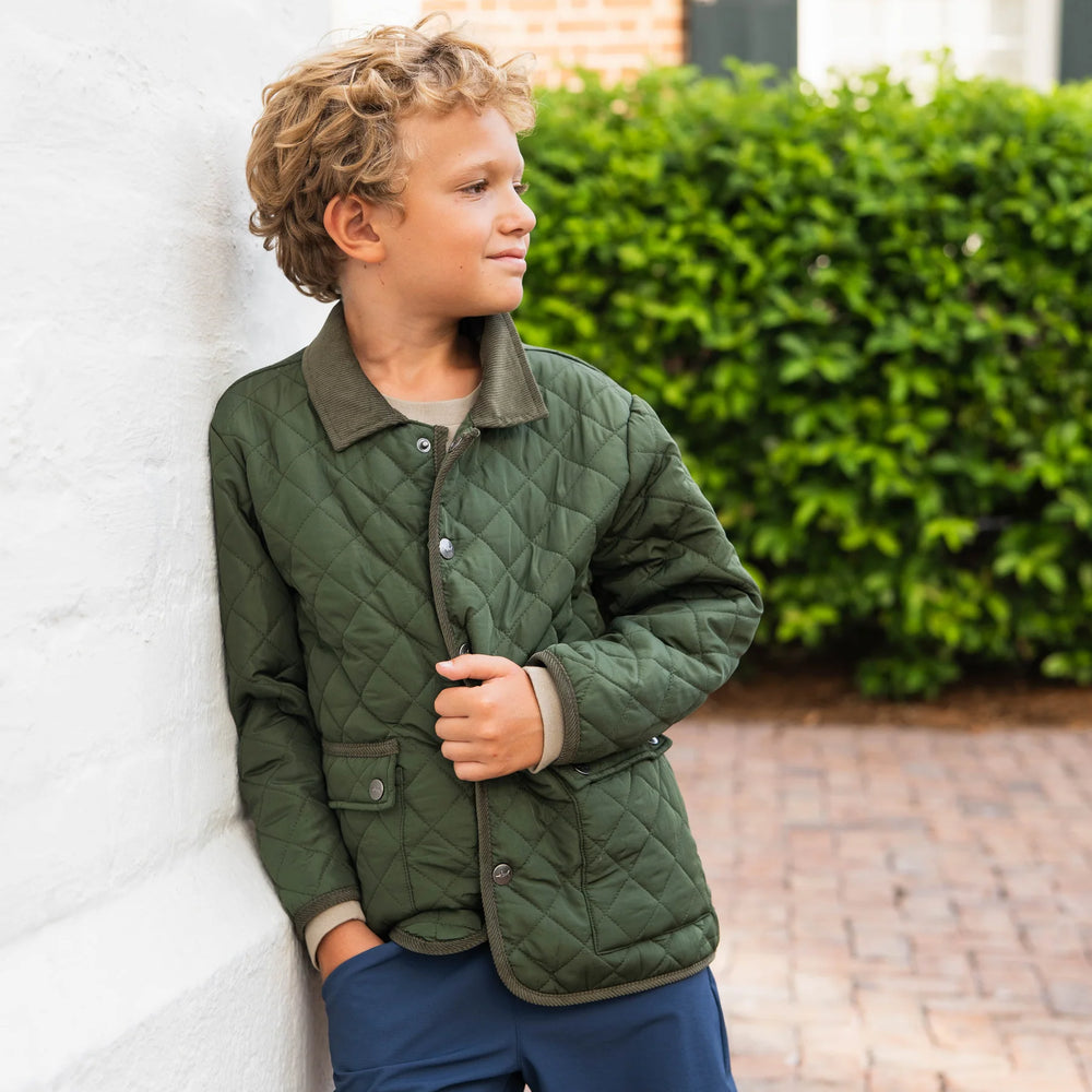 Young boy wearing Boys Derby Field Jacket in green paired with denim, leaning against outdoor wall, showcasing casual fall style and versatile layering.