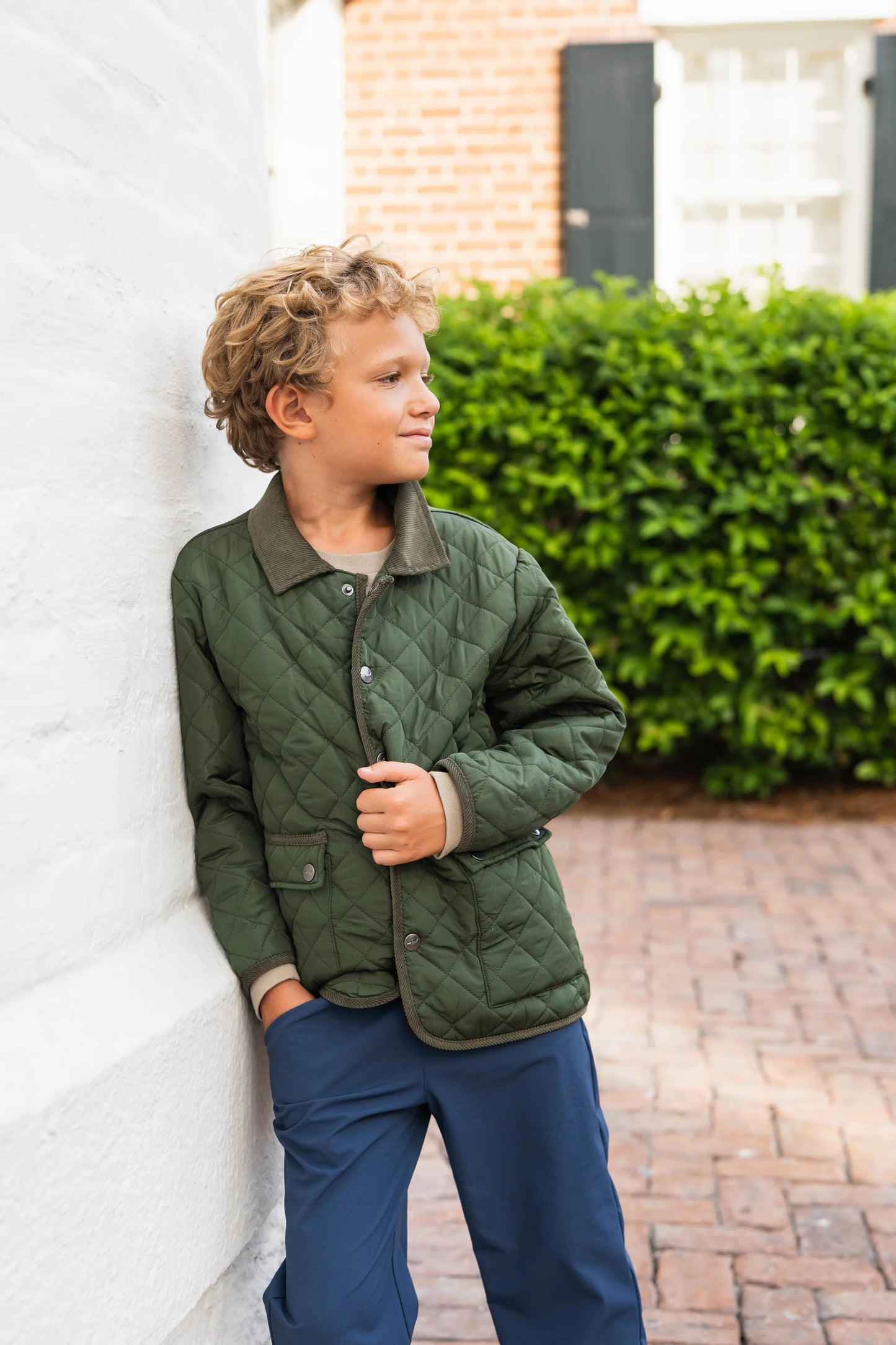 Young boy wearing Boys Derby Field Jacket in green paired with denim, leaning against outdoor wall, showcasing casual fall style and versatile layering.