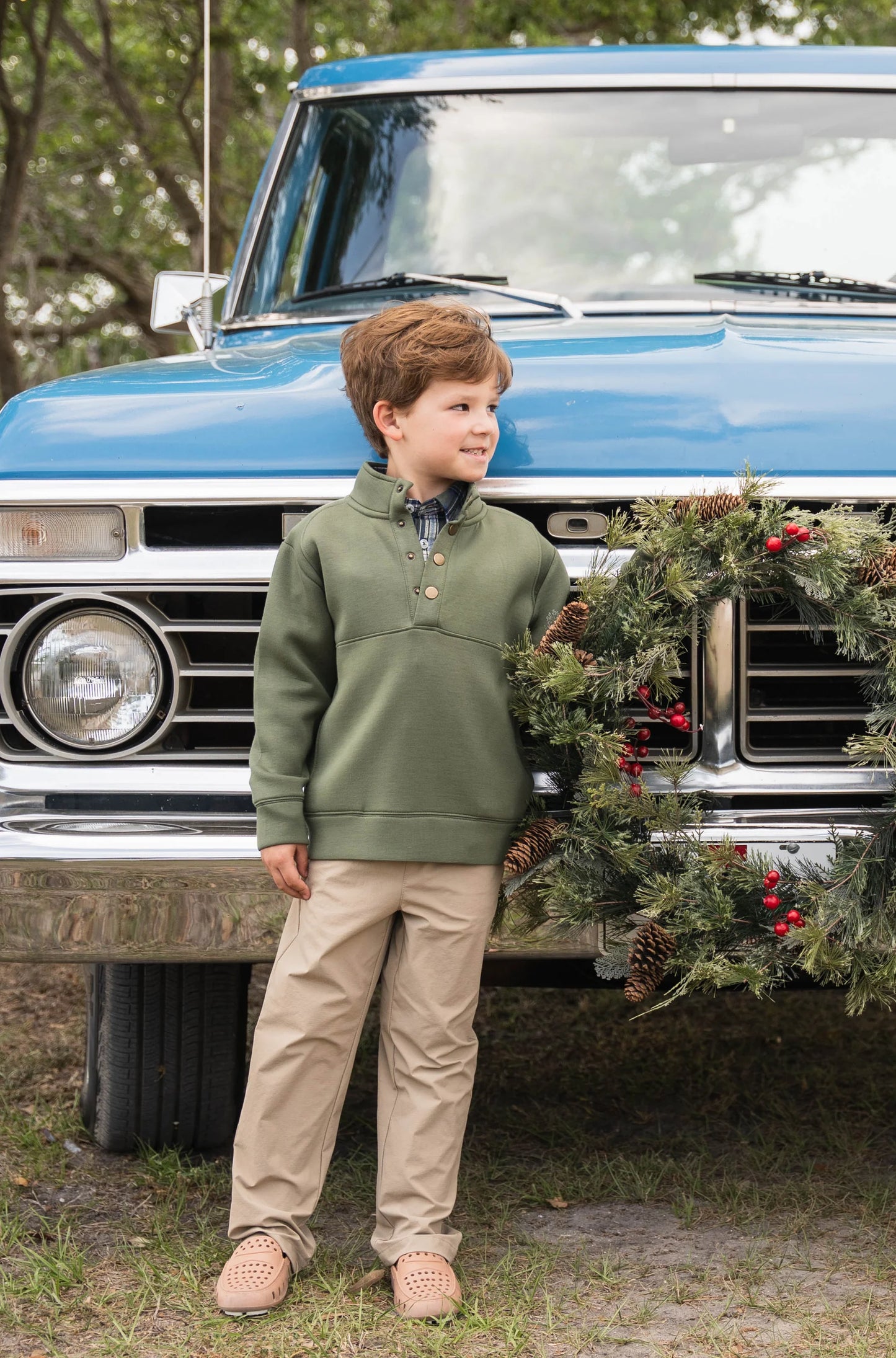 Boy wearing green Pierce Performance Pullover paired with performance pants, standing in front of vintage truck with holiday wreath, showcasing classic fall style.