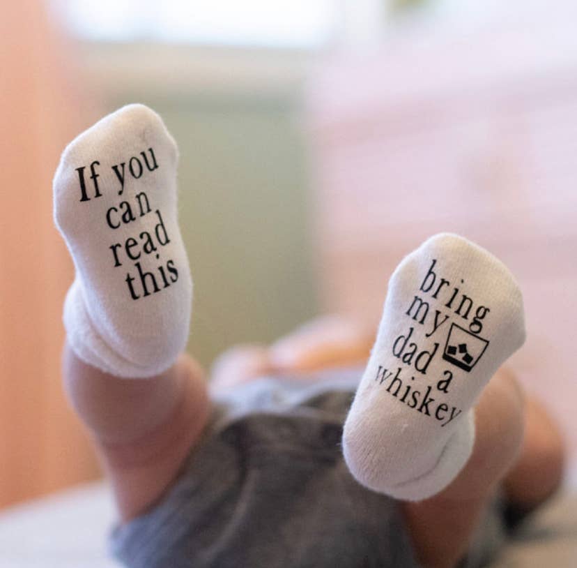 Close-up of baby socks featuring the “Bring My Dad Whiskey” message in bold text.