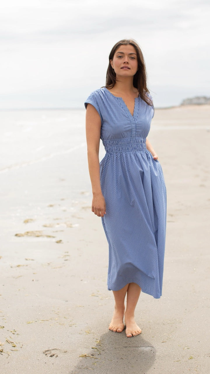 Model wearing the Clara Dress in blue and white pinstripe walking gracefully on the beach, showcasing the flowing half-circle skirt and fitted waist.