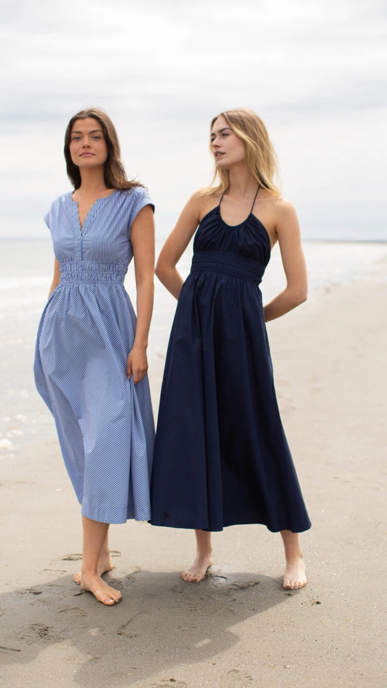 
                      
                        Two models walking together on the beach, one wearing the Clara Dress in blue and white pinstripe and the other in a navy Marea dress, both styled for a coastal look.
                      
                    