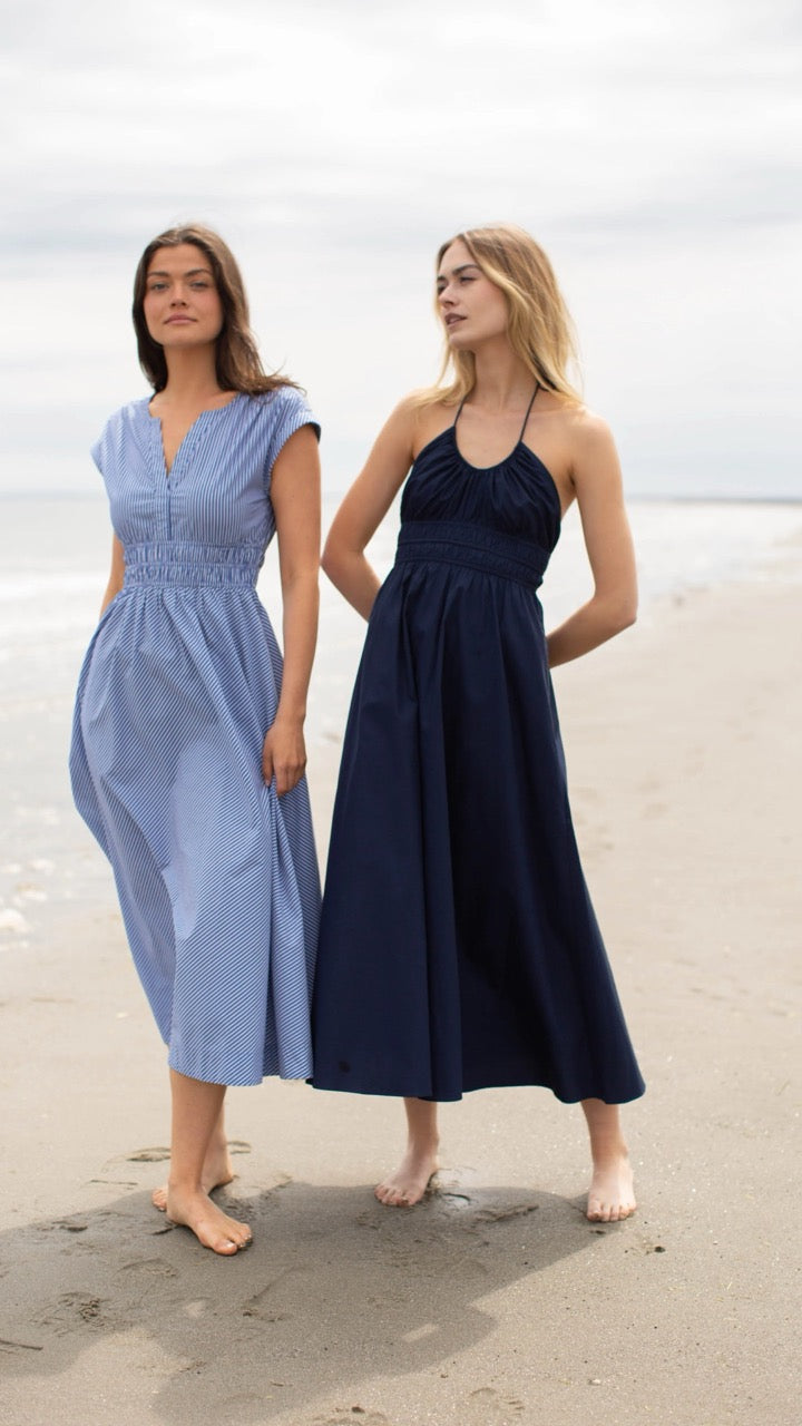Two models walking together on the beach, one wearing the Clara Dress in blue and white pinstripe and the other in a navy Marea dress, both styled for a coastal look.