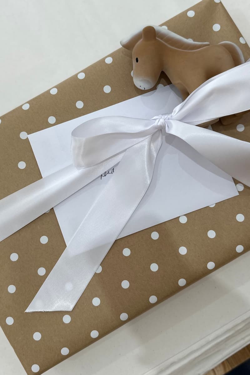 A gift box wrapped in brown kraft paper with white polka dots, tied with a large white satin ribbon and a white card, with a small wooden horse toy on top.