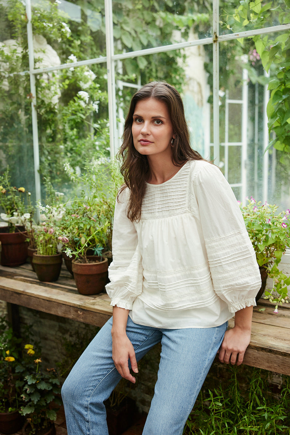 Model wearing the white Dawn Blouse in soft cotton voile, resting in a greenhouse styled casually with denim for a relaxed, feminine look.