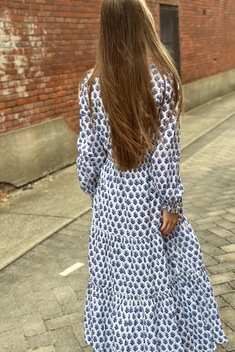 Woman in a long hand-blocked, floral Dottie dress in navy, blue & white walking on a sidewalk with a brick wall in the background
