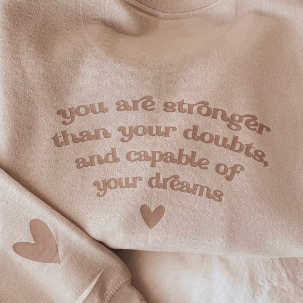 
                      
                        Neutral sand-colored sweatshirt with the inspirational message “You are stronger than your doubts, and capable of your dreams,” laid flat for full design view.
                      
                    