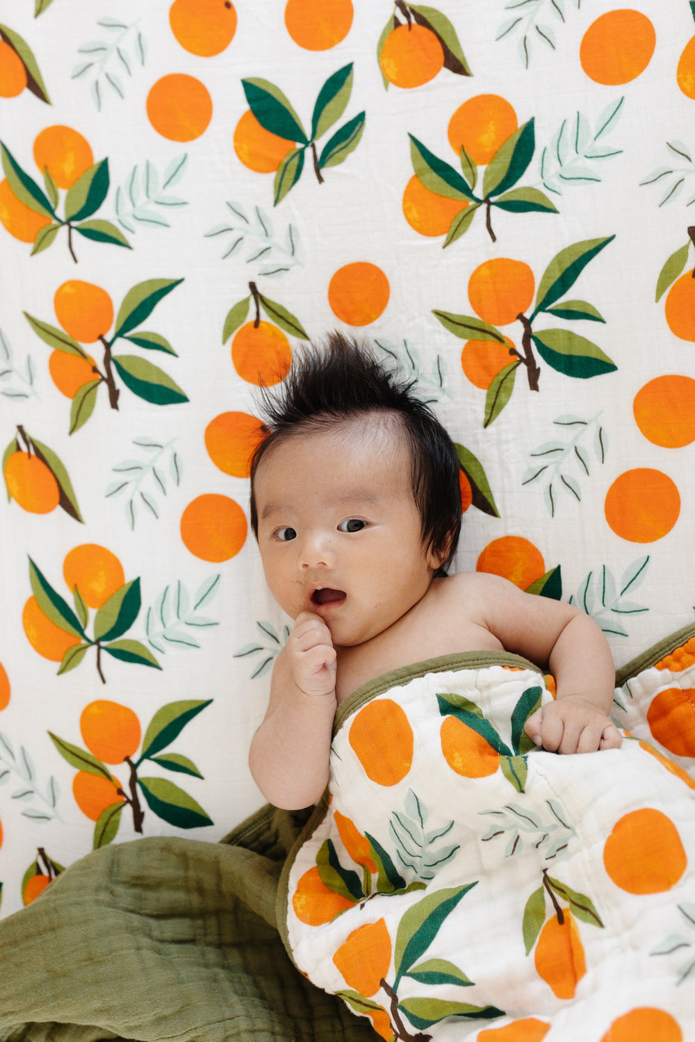 Baby wrapped in Fresh Clementine Quilt, laying on matching crib sheet and looking up at camera, showcasing ultra-soft 100% cotton muslin.