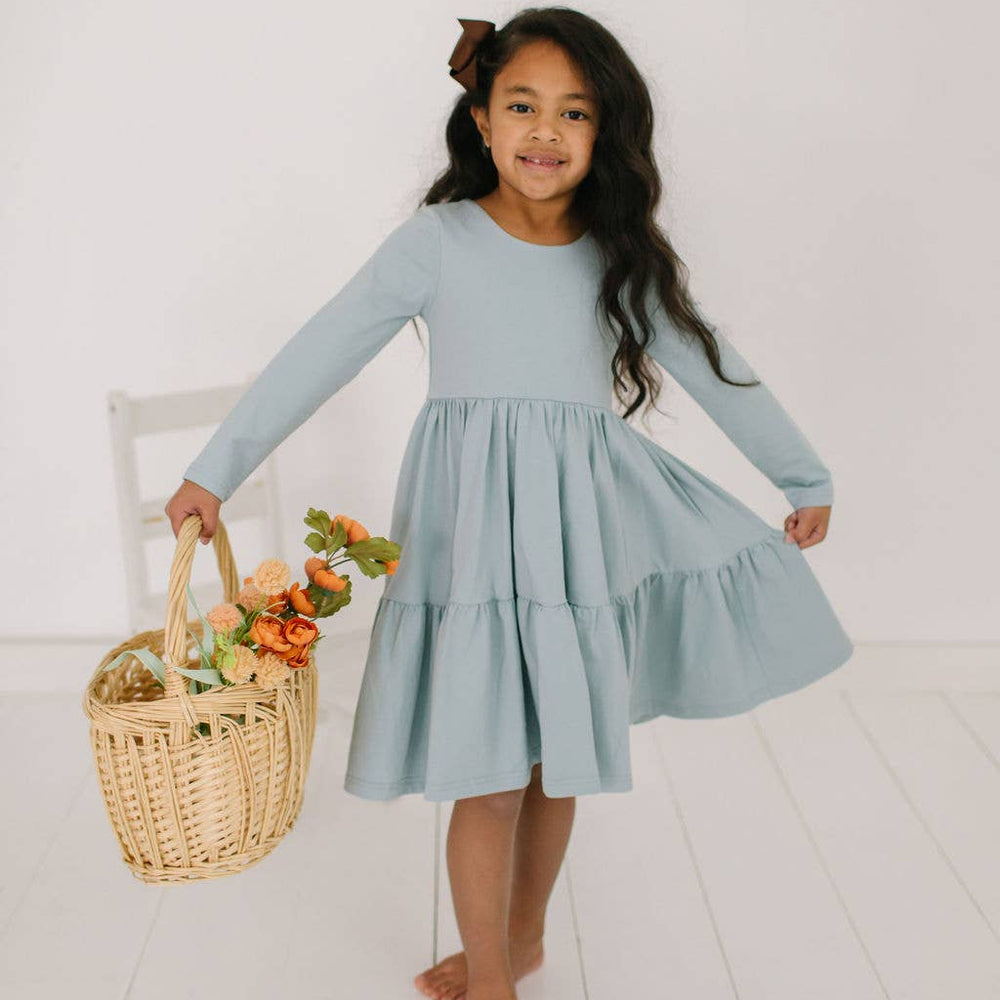 Toddler model holding out the skirt of the Gwendolyn Ruffle Dress in dusty blue to show its full twirl-ready design.