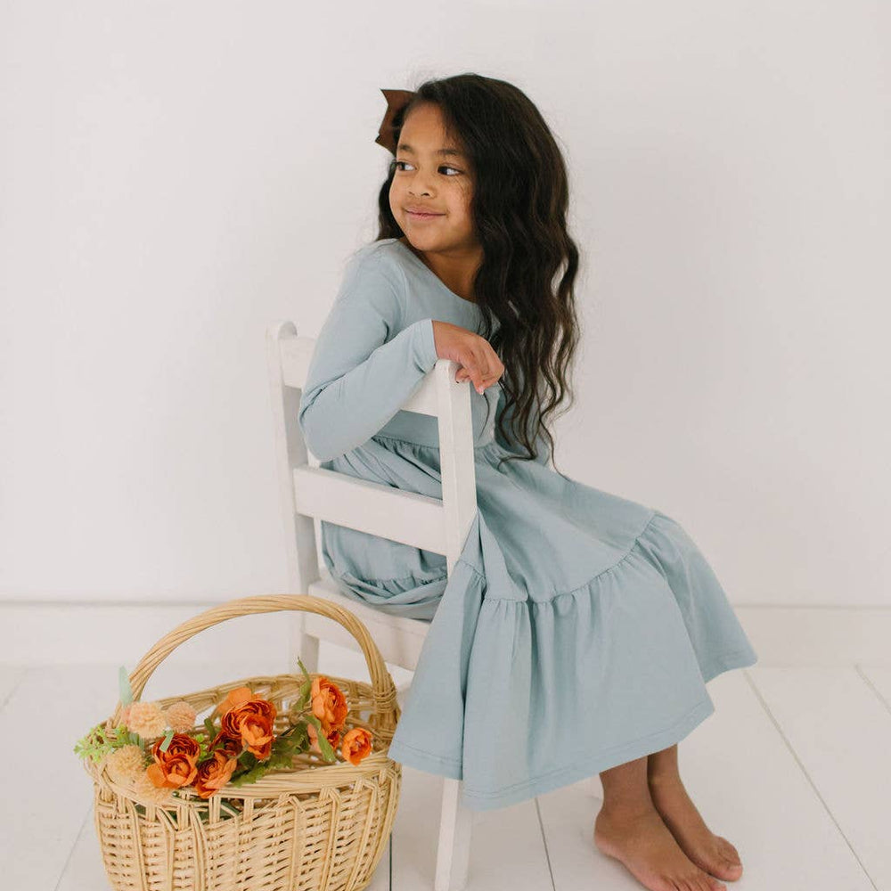 
                      
                        Toddler model sitting in the Gwendolyn Ruffle Dress in dusty blue, showing soft fabric and elegant ruffle accents.
                      
                    