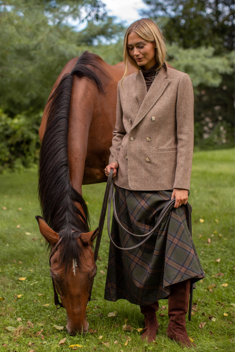 A woman standing outdoors in a grassy field next to a brown horse, wearing a beige double-breasted blazer, a green plaid skirt, and brown boots, holding the horse's reins. Skirt available at Grace & Haven.