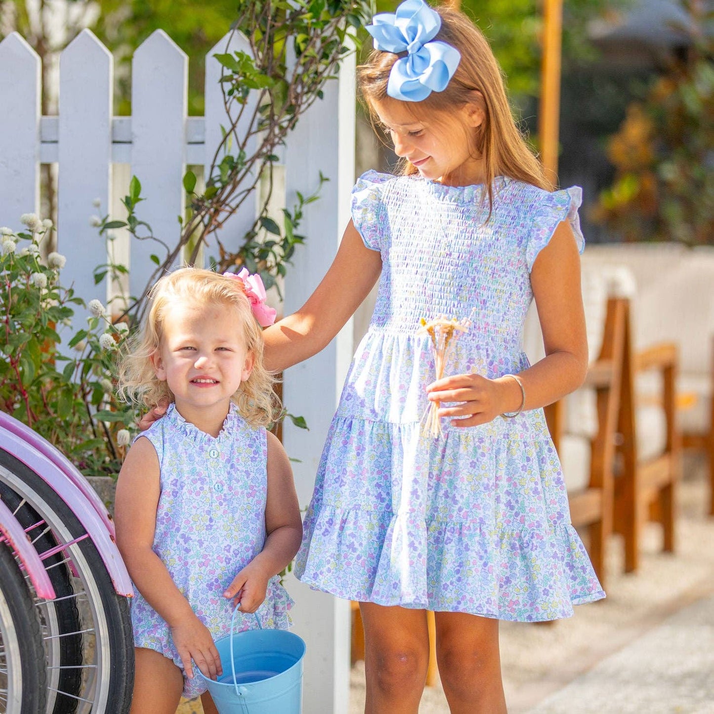 Front view of siblings in matching Kiawah outfits—older girl in the smocked twirl dress and younger sister in the coordinating ruffle bubble, styled for summer.