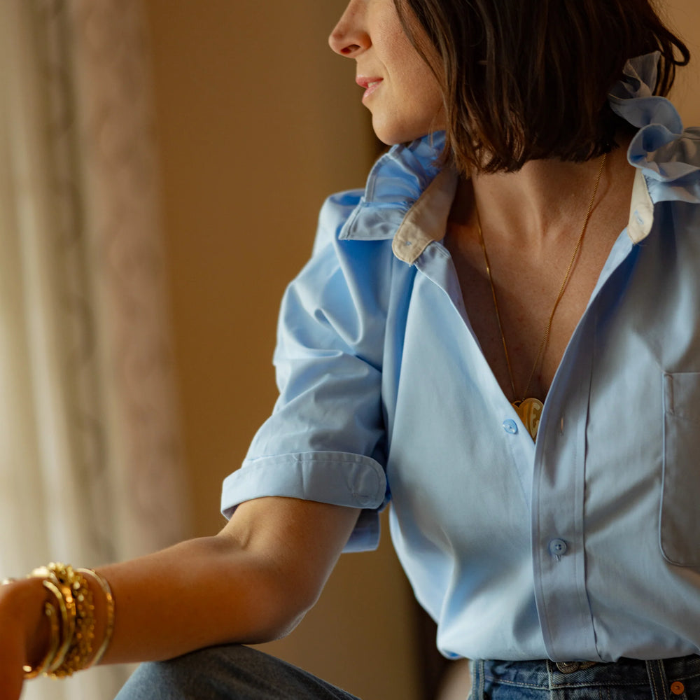 Designer Kate wearing KMJ Bonita light blue shirt with ruffle collar styled with casual shorts in a bright, peaceful hotel room in California.
