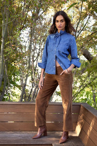 A woman stands outdoors on a wooden deck wearing a blue denim shirt with rolled sleeves, brown corduroy pants, and brown ankle boots, with trees in the background.
