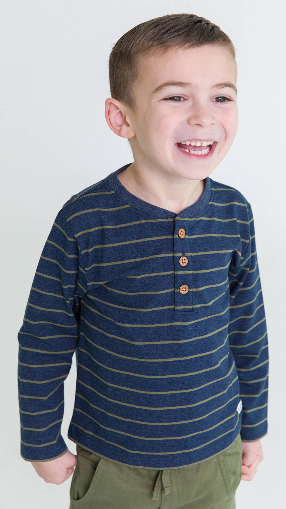 Young boy wearing the Boys Dusty Olive Stripe Knit Long Sleeve Henley Tee, smiling in a studio setting.