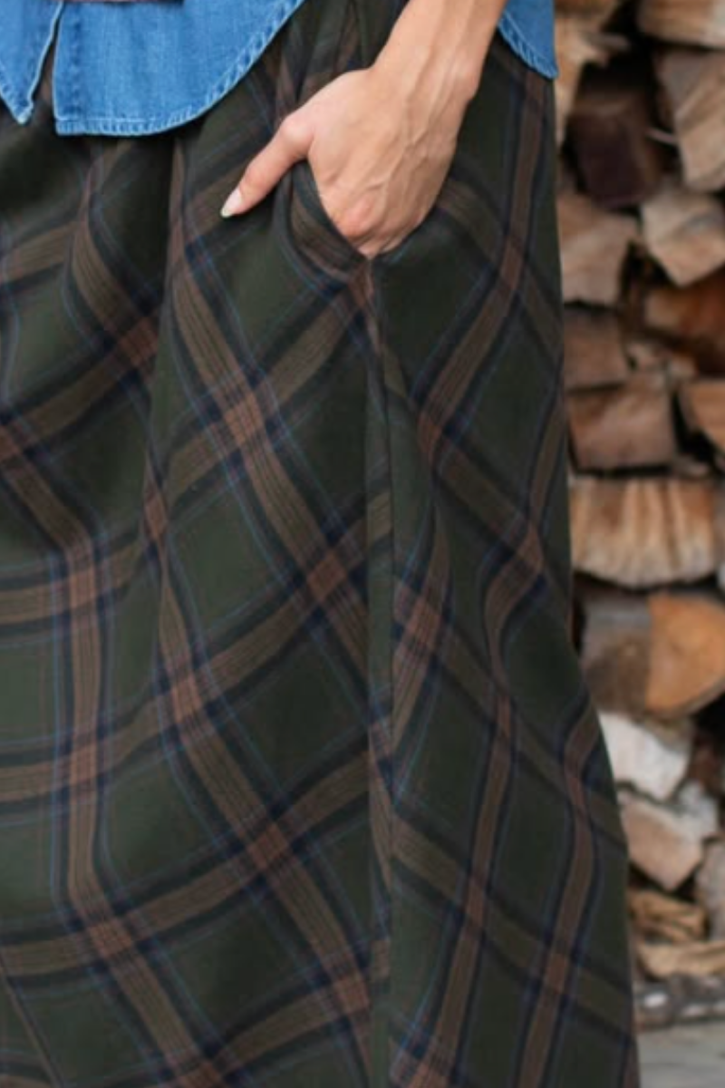 Close-up of a person wearing a dark green plaid skirt with brown and blue lines part of the Marea Fall Collection, holding a pocket, with a denim shirt untucked and a stack of firewood in the background. Available at Grace & Haven in Essex, CT or online at graceandhaven.co.