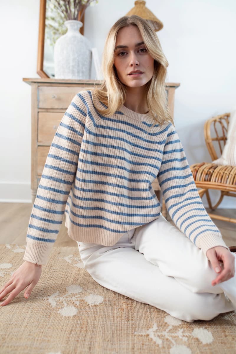 Woman wearing The Marea Sweater in Ivory Atlantic stripe sitting on a rug in a room with wooden furniture in Fairfield, CT. Sweater is available as part of the Fall & Winter Collection at Grace & Haven.