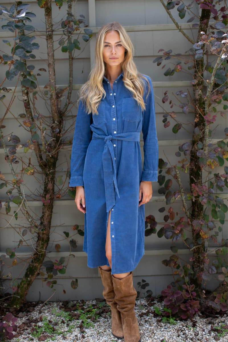 A woman stands outdoors in front of a gray house wall and leafy vines, wearing a blue corduroy shirt dress with a tie waist and brown suede boots. Marea fall dress available at Grace & Haven online at graceandhaven.co.