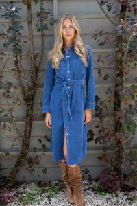 A woman stands outdoors in front of a gray house wall and leafy vines, wearing a blue corduroy shirt dress with a tie waist and brown suede boots. Marea fall dress available at Grace & Haven online at graceandhaven.co.