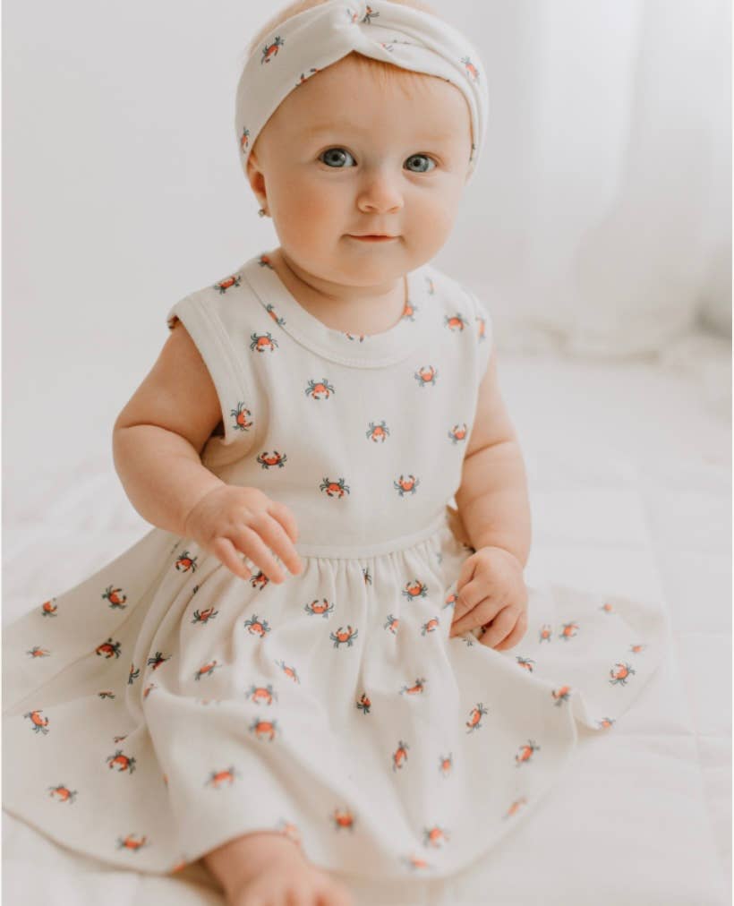 Toddler girl sitting in Organic Cotton Crab Twirl Dress with matching headband, smiling sweetly