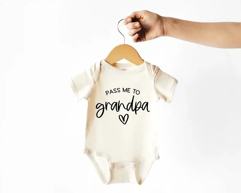 Close-up of “Pass Me to Grandpa” baby bodysuit in soft natural beige with bold printed message.