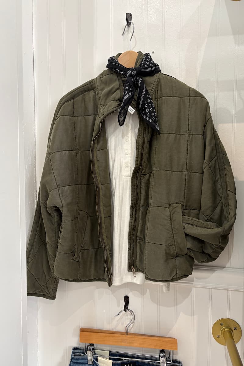 An olive green quilted jacket hanging on a wooden hanger against a white paneled wall at Grace & Haven in Essex, CT, styled with a black patterned neck scarf and a white Marea pointelle henley shirt underneath.