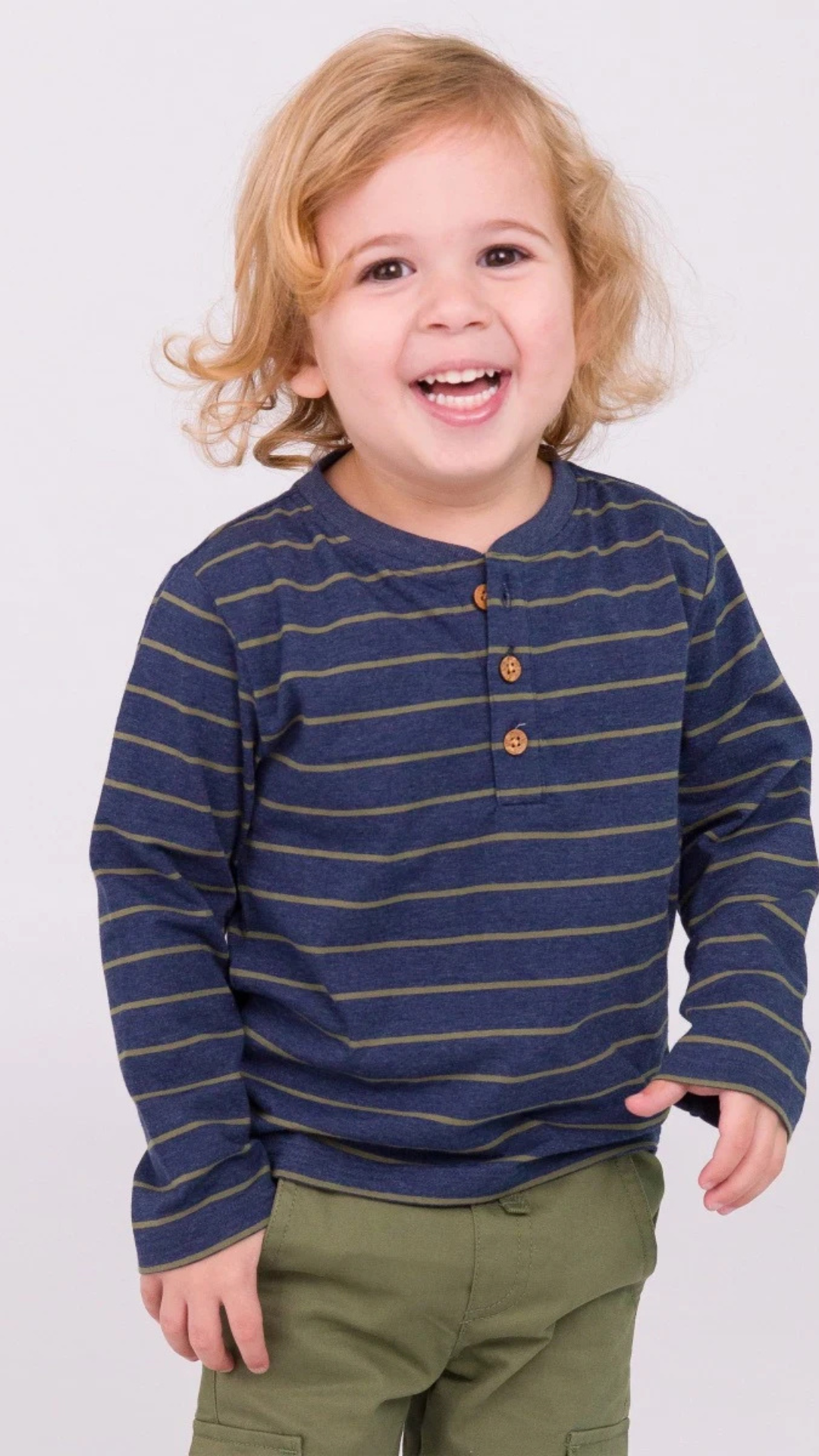 Happy little boy wearing the Boys Dusty Olive Stripe Knit Long Sleeve Henley Tee, showcasing its stylish design in a bright studio.